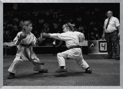 A black and white photograph of two young girls in a karate competition. They are wearing white gis, black belts, and boxing gloves. One girl is in the middle of throwing a punch at her opponent. In the background, a referee stands watching the match, and spectators are blurred in the stands. The image is presented within a silver-colored frame. Print