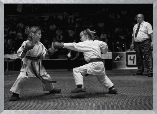 A black and white photograph of two young girls in a karate competition. They are wearing white gis, black belts, and boxing gloves. One girl is in the middle of throwing a punch at her opponent. In the background, a referee stands watching the match, and spectators are blurred in the stands. The image is presented within a silver-colored frame. Print