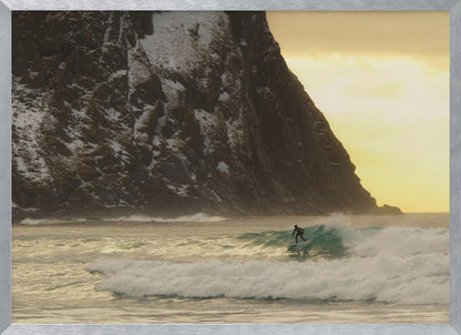 A framed photograph captures a lone surfer in a black wetsuit riding a turquoise wave. In the background, a massive, steep, snow-dusted mountain looms over the cold sea under a pale yellow, overcast sky. Print