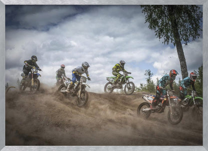 A dynamic action shot of several motocross riders racing up a dirt hill, kicking up a large cloud of dust. The riders are in full gear on colorful bikes, with a dramatic cloudy sky and a tall tree in the background. Print