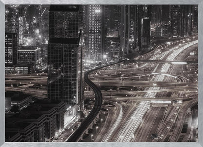 A dynamic black and white long-exposure photograph of a sprawling metropolis at night, viewed from above. Gleaming skyscrapers fill the background, while a complex interchange of highways with streaks of light from traffic dominates the foreground, with an elevated train line weaving through the city. Wall Art