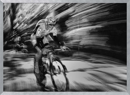 A dramatic black and white action shot of a professional cyclist racing. The cyclist is in focus, wearing a helmet and sunglasses, while the background is a streaky motion blur, creating a powerful sense of speed and movement. The image is enclosed in a silver frame. Wall Art