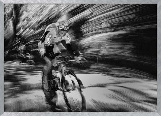 A dramatic black and white action shot of a professional cyclist racing. The cyclist is in focus, wearing a helmet and sunglasses, while the background is a streaky motion blur, creating a powerful sense of speed and movement. The image is enclosed in a silver frame. Wall Art