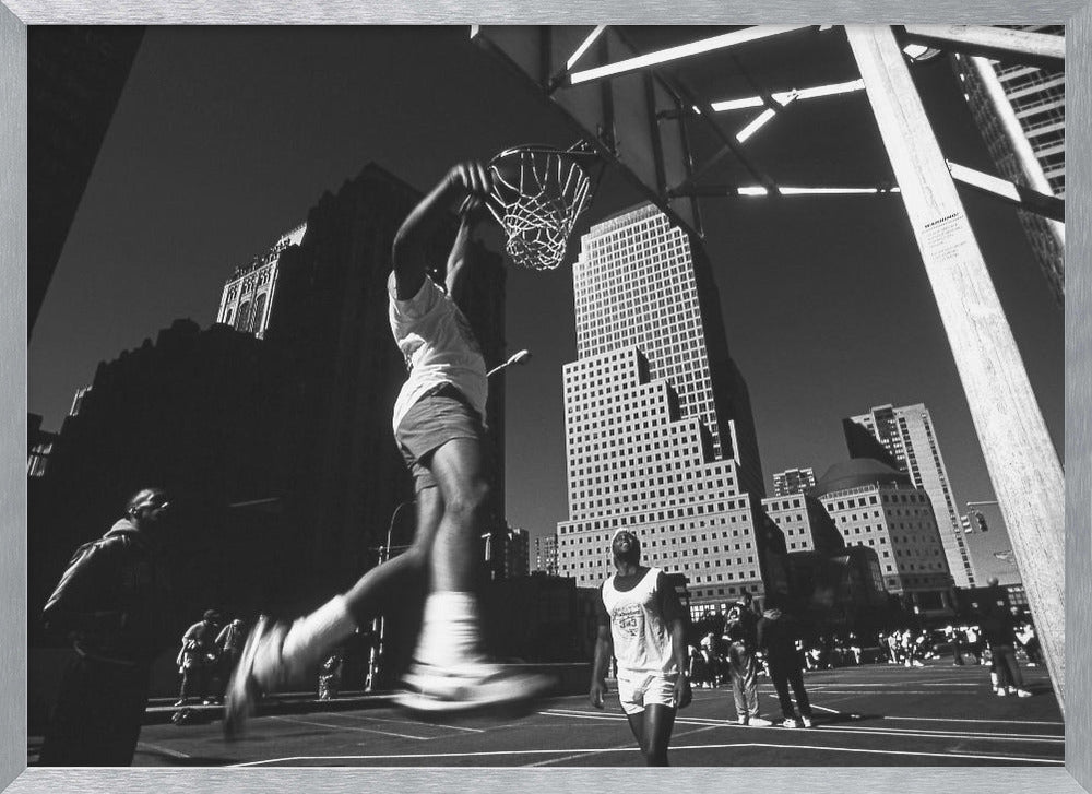 A low-angle, black and white action shot of a person dunking a basketball on an outdoor court, with tall city skyscrapers dominating the background under a clear sky. Print