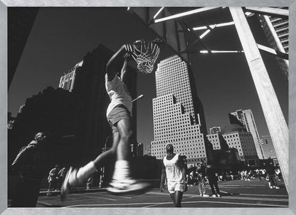A low-angle, black and white action shot of a person dunking a basketball on an outdoor court, with tall city skyscrapers dominating the background under a clear sky. Print