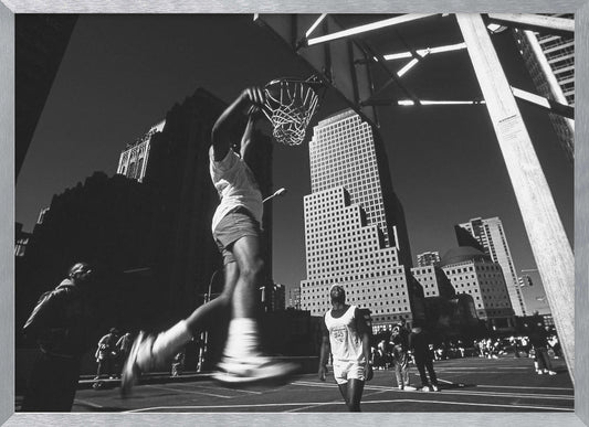 A low-angle, black and white action shot of a person dunking a basketball on an outdoor court, with tall city skyscrapers dominating the background under a clear sky. Print