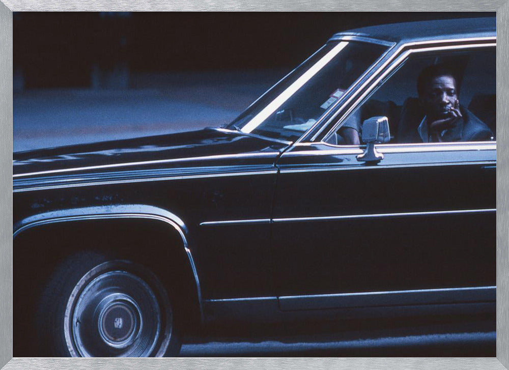 A moody, blue-toned photograph of a Black man sitting pensively in the driver's seat of a dark vintage sedan at night, framed in silver. Poster