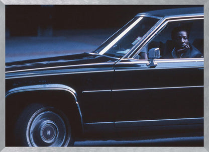 A moody, blue-toned photograph of a Black man sitting pensively in the driver's seat of a dark vintage sedan at night, framed in silver. Poster