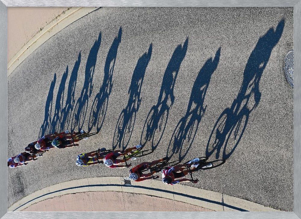 An aerial, high-angle photograph of a line of cyclists racing on a curved asphalt road. The bright sun casts long, dark, dramatic shadows of the riders and their bicycles, creating a striking diagonal pattern across the pavement. The image is framed in silver. Print