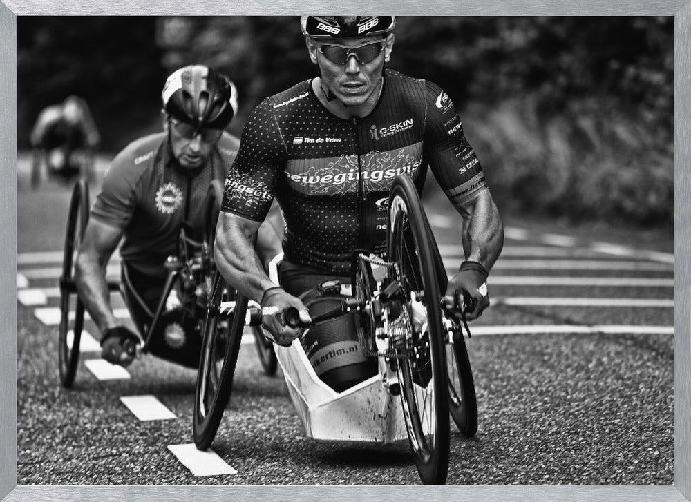 A powerful black and white photograph of two male athletes competing in a wheelchair race. The focused athlete in the foreground shows intense determination on his face, his muscular arms propelling the specialized racing wheelchair forward on an asphalt road. A second racer is visible in the background. Decor