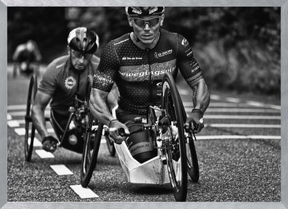 A powerful black and white photograph of two male athletes competing in a wheelchair race. The focused athlete in the foreground shows intense determination on his face, his muscular arms propelling the specialized racing wheelchair forward on an asphalt road. A second racer is visible in the background. Decor