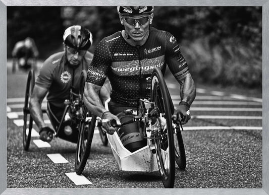 A powerful black and white photograph of two male athletes competing in a wheelchair race. The focused athlete in the foreground shows intense determination on his face, his muscular arms propelling the specialized racing wheelchair forward on an asphalt road. A second racer is visible in the background. Decor