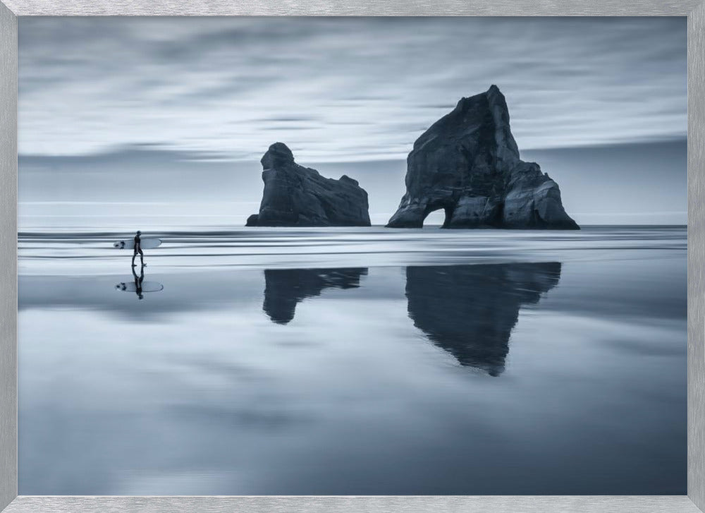A moody, blue-toned monochromatic photograph of a lone surfer walking on a wet, reflective beach. Two large rock formations stand in the calm ocean in the background, their reflections mirrored in the water. The sky is overcast, creating a serene and atmospheric scene, all enclosed in a silver frame. Decor
