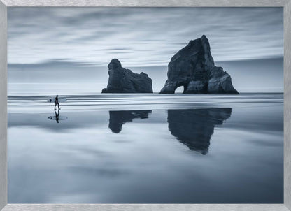 A moody, blue-toned monochromatic photograph of a lone surfer walking on a wet, reflective beach. Two large rock formations stand in the calm ocean in the background, their reflections mirrored in the water. The sky is overcast, creating a serene and atmospheric scene, all enclosed in a silver frame. Decor