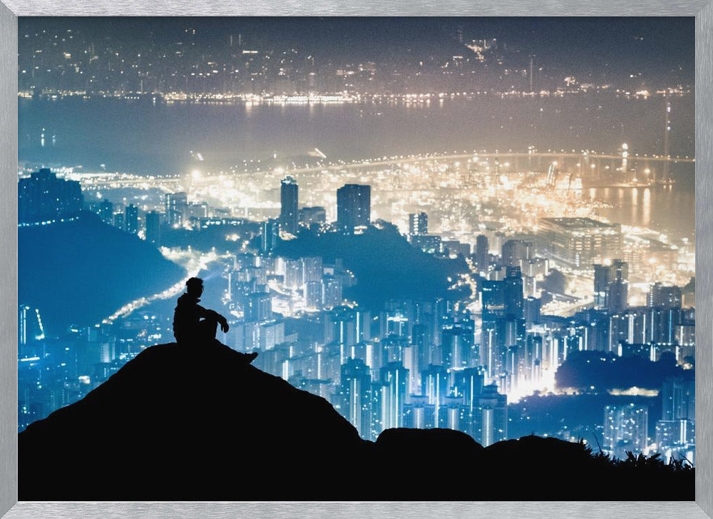 A silhouette of a person sitting on a hilltop, gazing out over a vast, illuminated city skyline at night. The city lights glow in shades of blue and gold against the dark landscape, with a body of water in the distance. Decor