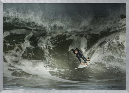 A dramatic overhead photograph of a surfer in a black wetsuit riding a white surfboard on a large, dark, foamy wave. The scene is captured in a moody, almost monochromatic tone and is presented within a silver picture frame. Artwork