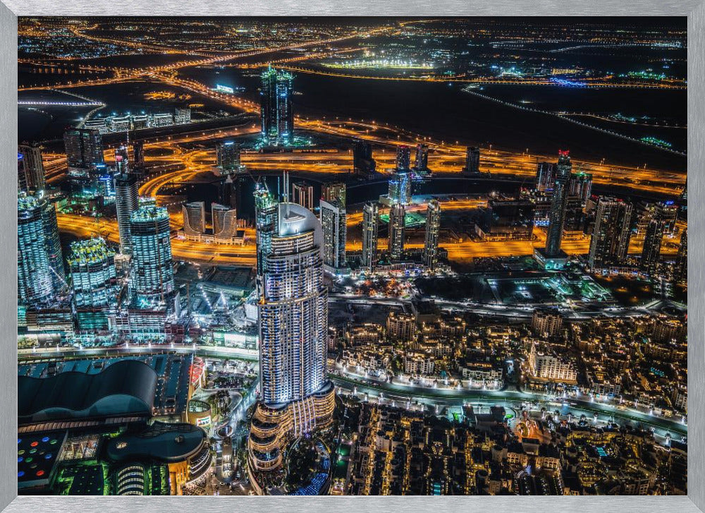 An aerial nighttime photograph of a sprawling modern city, possibly Dubai, with brightly lit skyscrapers and a complex network of illuminated highways creating a vibrant contrast against the dark landscape, all enclosed in a silver frame. Decor