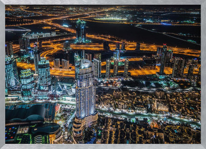 An aerial nighttime photograph of a sprawling modern city, possibly Dubai, with brightly lit skyscrapers and a complex network of illuminated highways creating a vibrant contrast against the dark landscape, all enclosed in a silver frame. Decor