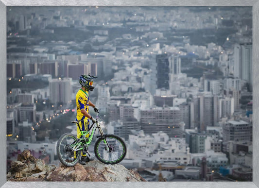 A mountain biker in a yellow and blue uniform stands with a green and white full-suspension bike on a rocky precipice, looking out over a vast, hazy cityscape that stretches into the distance. Print