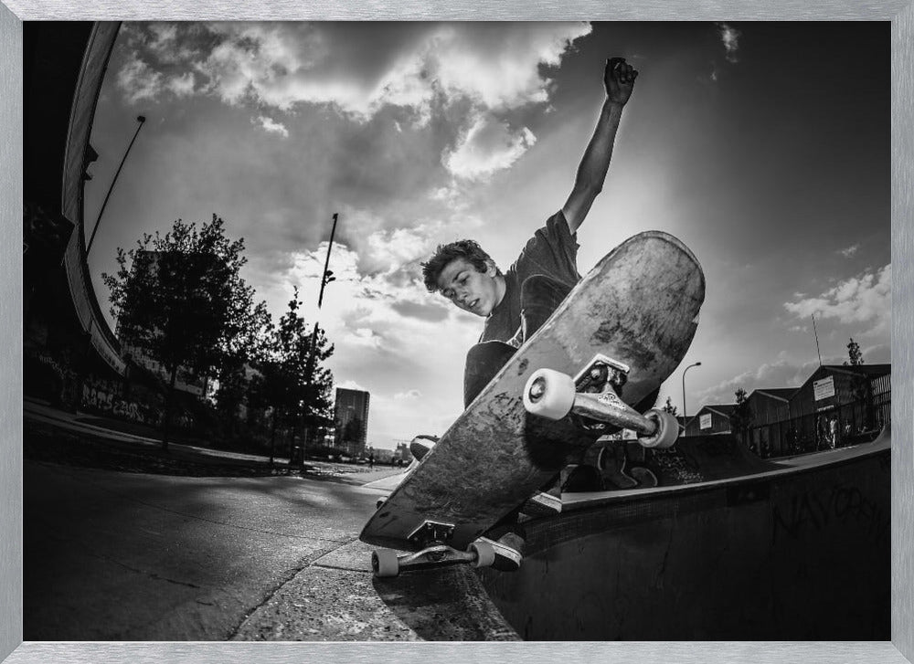 A dynamic, low-angle, black and white photograph of a young man skateboarding. He is balancing on the edge of a ramp with his skateboard angled up towards the viewer. He has one arm raised in the air with a clenched fist. The shot is taken with a fisheye lens, curving the horizon against a dramatic cloudy sky in an urban setting. The image is presented within a silver frame. Decor