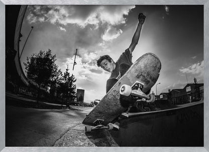 A dynamic, low-angle, black and white photograph of a young man skateboarding. He is balancing on the edge of a ramp with his skateboard angled up towards the viewer. He has one arm raised in the air with a clenched fist. The shot is taken with a fisheye lens, curving the horizon against a dramatic cloudy sky in an urban setting. The image is presented within a silver frame. Decor