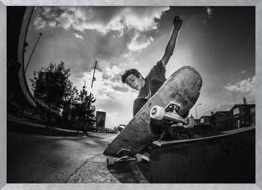 A dynamic, low-angle, black and white photograph of a young man skateboarding. He is balancing on the edge of a ramp with his skateboard angled up towards the viewer. He has one arm raised in the air with a clenched fist. The shot is taken with a fisheye lens, curving the horizon against a dramatic cloudy sky in an urban setting. The image is presented within a silver frame. Decor