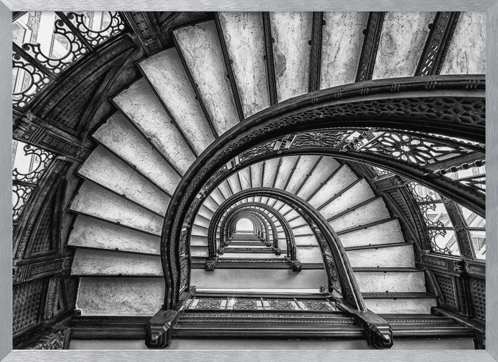 A dramatic black and white photograph looking up from the bottom of an ornate spiral staircase. The dark, intricate ironwork of the railings contrasts with the light stone steps, creating a mesmerizing, repeating pattern that spirals towards a bright light at the top. The image is presented within a silver-colored frame. Decor