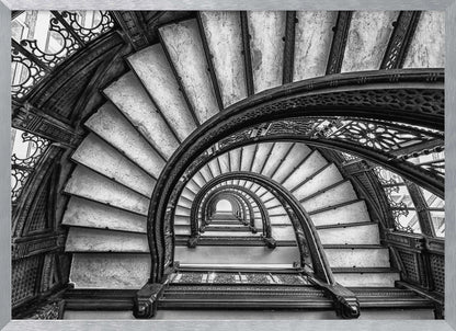 A dramatic black and white photograph looking up from the bottom of an ornate spiral staircase. The dark, intricate ironwork of the railings contrasts with the light stone steps, creating a mesmerizing, repeating pattern that spirals towards a bright light at the top. The image is presented within a silver-colored frame. Decor