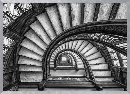 A dramatic black and white photograph looking up from the bottom of an ornate spiral staircase. The dark, intricate ironwork of the railings contrasts with the light stone steps, creating a mesmerizing, repeating pattern that spirals towards a bright light at the top. The image is presented within a silver-colored frame. Decor