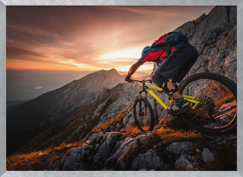 A mountain biker in full gear rides a yellow bike down a narrow, rocky mountain ridge at sunset. The sky is filled with the warm colors of the setting sun, which illuminates the distant mountain peaks. Artwork
