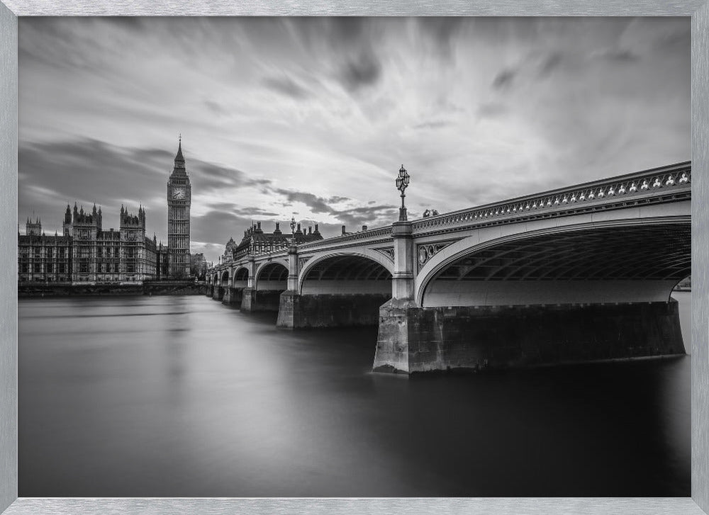 A dramatic black and white long-exposure photograph of London, featuring the smooth River Thames in the foreground, Westminster Bridge on the right, and the iconic Big Ben and Houses of Parliament on the left under a cloudy, streaked sky. The image is presented in a silver frame. Print