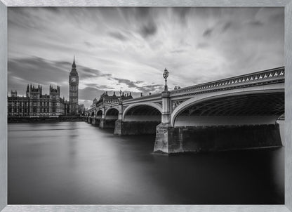 A dramatic black and white long-exposure photograph of London, featuring the smooth River Thames in the foreground, Westminster Bridge on the right, and the iconic Big Ben and Houses of Parliament on the left under a cloudy, streaked sky. The image is presented in a silver frame. Print