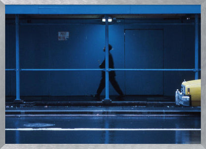 A moody, grainy photograph dominated by shades of blue, capturing the blurred silhouette of a person walking under a covered city sidewalk at night. The wet street in the foreground reflects light, and the yellow cab of a truck is partially visible on the right. Artwork