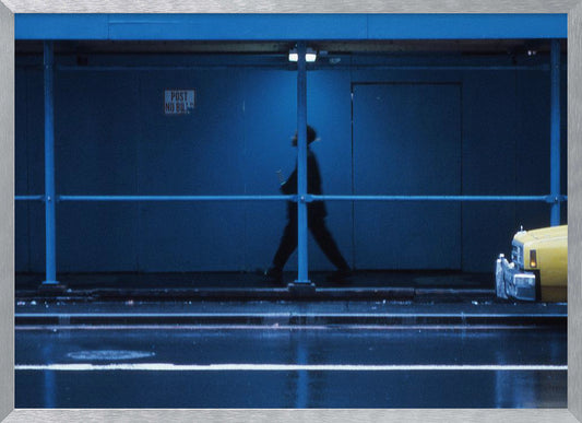 A moody, grainy photograph dominated by shades of blue, capturing the blurred silhouette of a person walking under a covered city sidewalk at night. The wet street in the foreground reflects light, and the yellow cab of a truck is partially visible on the right. Artwork