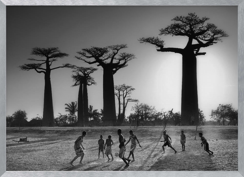 A framed, black and white photograph capturing a vibrant scene of several children playing soccer on a dirt field. In the background, the iconic silhouettes of large Baobab trees stand tall against a bright, hazy sun, casting long shadows of the players across the ground. Artwork