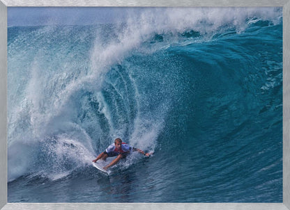 A dramatic action shot of a professional surfer skillfully navigating the inside of a massive, curling blue ocean wave, surrounded by white sea spray, all enclosed in a silver frame. Artwork
