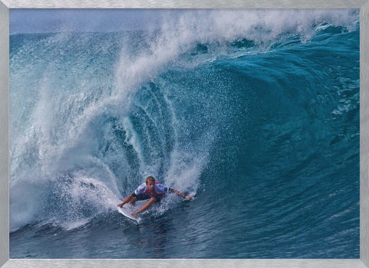 A dramatic action shot of a professional surfer skillfully navigating the inside of a massive, curling blue ocean wave, surrounded by white sea spray, all enclosed in a silver frame. Artwork