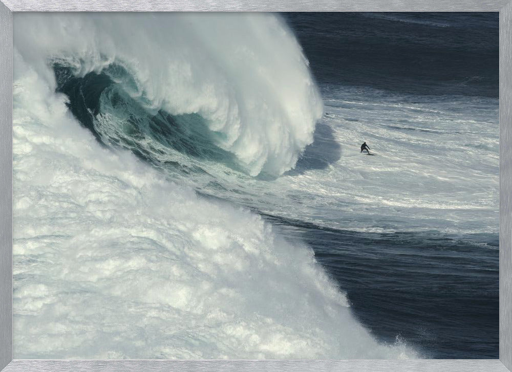 A framed aerial photograph captures a dramatic ocean scene where a lone surfer on a surfboard is dwarfed by an immense, powerful wave cresting nearby, conveying a sense of scale and adventure. Artwork