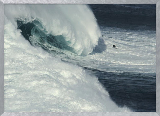 A framed aerial photograph captures a dramatic ocean scene where a lone surfer on a surfboard is dwarfed by an immense, powerful wave cresting nearby, conveying a sense of scale and adventure. Artwork