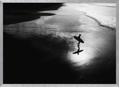 A high-contrast, black and white photograph of a silhouetted surfer carrying a surfboard and walking along the wet, reflective sand at the water's edge. Decor