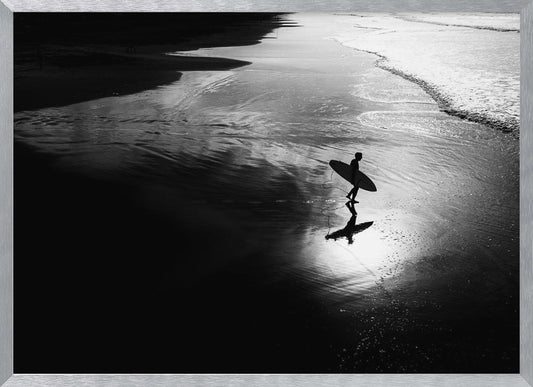 A high-contrast, black and white photograph of a silhouetted surfer carrying a surfboard and walking along the wet, reflective sand at the water's edge. Decor