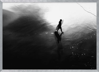 A high-contrast, black and white photograph of a surfer silhouetted against the bright reflection of the sun on the water. The surfer is carrying a surfboard and walking along the wet sand, leaving footprints behind. Their long shadow is cast in front of them on the dark, glistening sand. Print