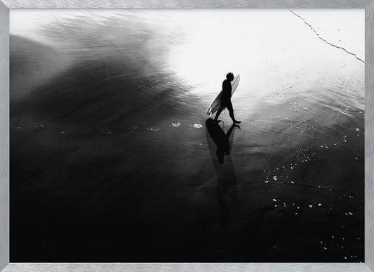 A high-contrast, black and white photograph of a surfer silhouetted against the bright reflection of the sun on the water. The surfer is carrying a surfboard and walking along the wet sand, leaving footprints behind. Their long shadow is cast in front of them on the dark, glistening sand. Print