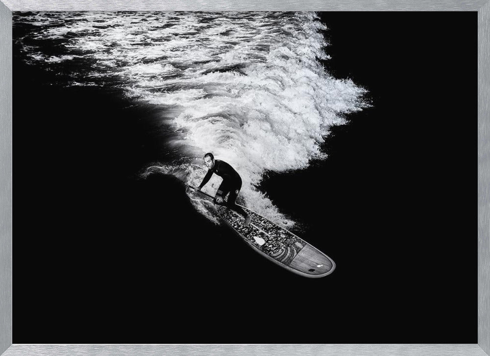 A dramatic black and white high-angle photograph of a man in a wetsuit paddleboarding on a wave, creating a stark white wake against the deep black water, all enclosed in a silver frame. Print