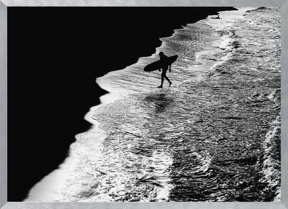 A high-contrast, black and white aerial photograph of a silhouetted surfer carrying a surfboard and walking along the shoreline. The image is divided diagonally, with the dark beach on the left and the white, foamy ocean waves on the right, creating a dramatic and graphic composition. The image is enclosed in a silver frame. Wall Art