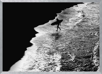 A high-contrast, black and white aerial photograph of a silhouetted surfer carrying a surfboard and walking along the shoreline. The image is divided diagonally, with the dark beach on the left and the white, foamy ocean waves on the right, creating a dramatic and graphic composition. The image is enclosed in a silver frame. Wall Art