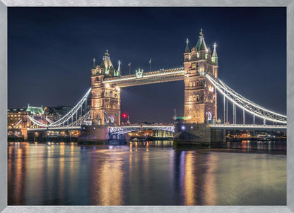 A stunning night photograph of London's iconic Tower Bridge, fully illuminated with golden and white lights that cast a shimmering reflection on the dark waters of the River Thames below, all set against a deep night sky and enclosed in a silver frame. Poster