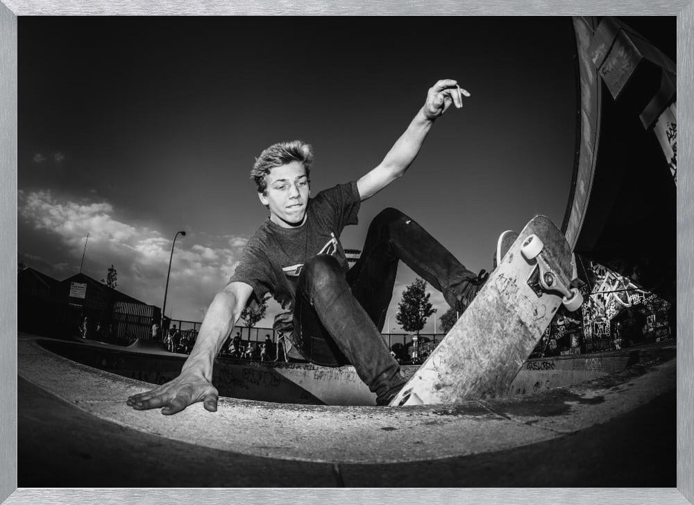 A dynamic, low-angle black and white action shot of a young skateboarder performing a trick in a concrete skatepark. He balances with one hand on the edge of the bowl, his body low to the ground as he carves along the curve, with his other arm raised in the air. Wall Art