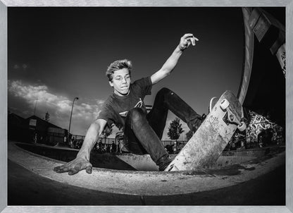 A dynamic, low-angle black and white action shot of a young skateboarder performing a trick in a concrete skatepark. He balances with one hand on the edge of the bowl, his body low to the ground as he carves along the curve, with his other arm raised in the air. Wall Art