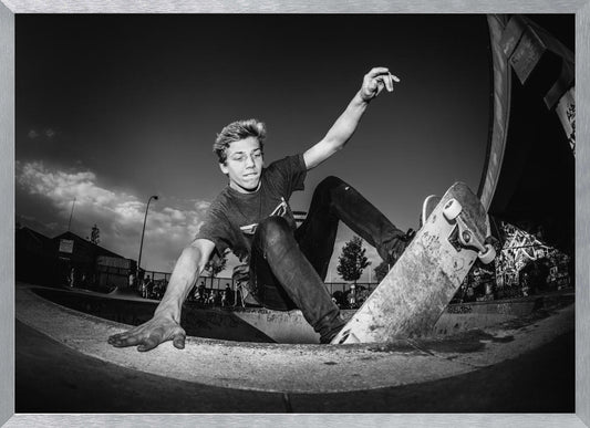 A dynamic, low-angle black and white action shot of a young skateboarder performing a trick in a concrete skatepark. He balances with one hand on the edge of the bowl, his body low to the ground as he carves along the curve, with his other arm raised in the air. Wall Art